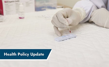 A gloved hand holding a dropper over a test kit with "Health Policy Update" text.