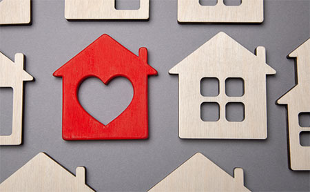 A red house-shaped cutout with a heart in the center, surrounded by various light wood-tone house silhouettes.