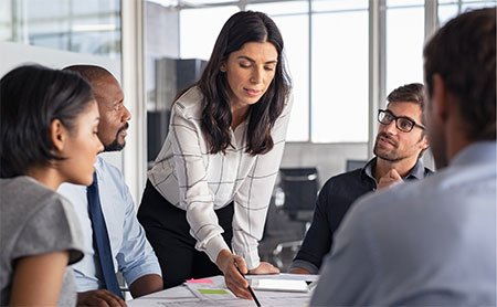 A group of individuals engaged in a meeting around a table, with one person standing and pointing at documents.